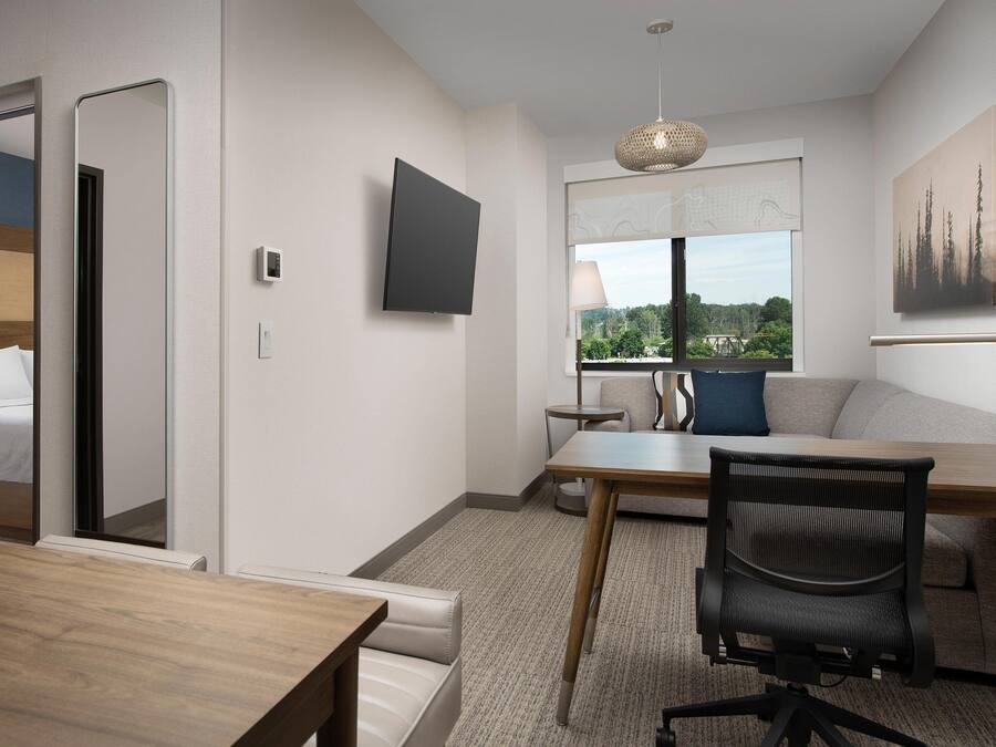 Una habitación de hotel moderna con una mesa de comedor de madera, una silla de oficina negra, un sofá, un televisor de pantalla plana montado en la pared y una ventana con vistas a zonas verdes.