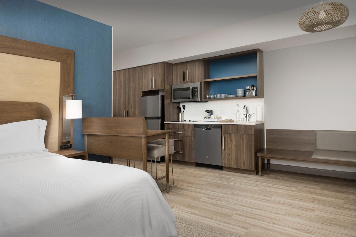 A modern hotel room with a bed, a desk, a kitchenette, and a seating area.