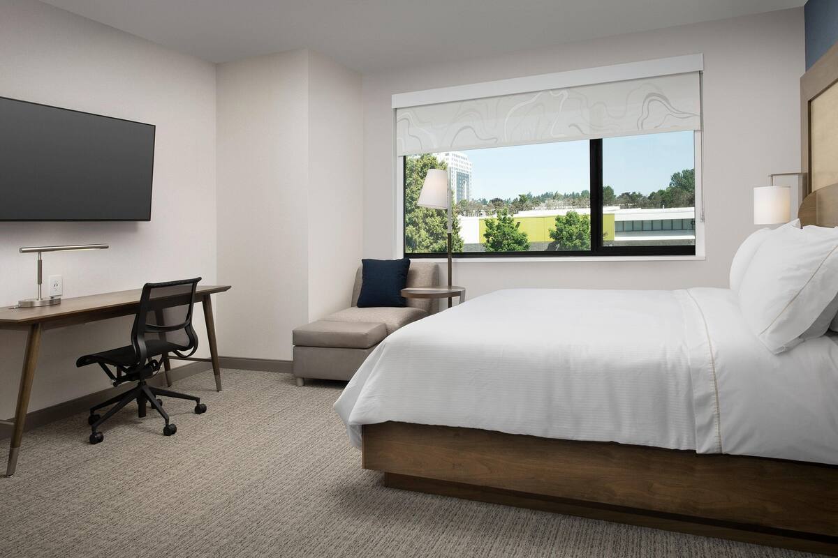 A hotel room with a bed, a desk with a chair, a TV, a window with a view of buildings, and a lamp.