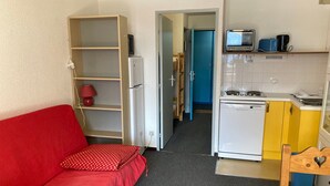 Un espace de vie compact comprenant un canapé rouge, une petite kitchenette et une porte donnant sur une autre pièce.