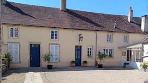 Un bâtiment à deux étages, avec un toit recouvert de tuiles, une porte bleue au centre et des plantes en pot.