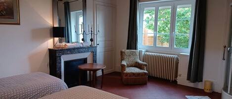 A bedroom with two beds, a fireplace, a window with a view of greenery, and a chair.