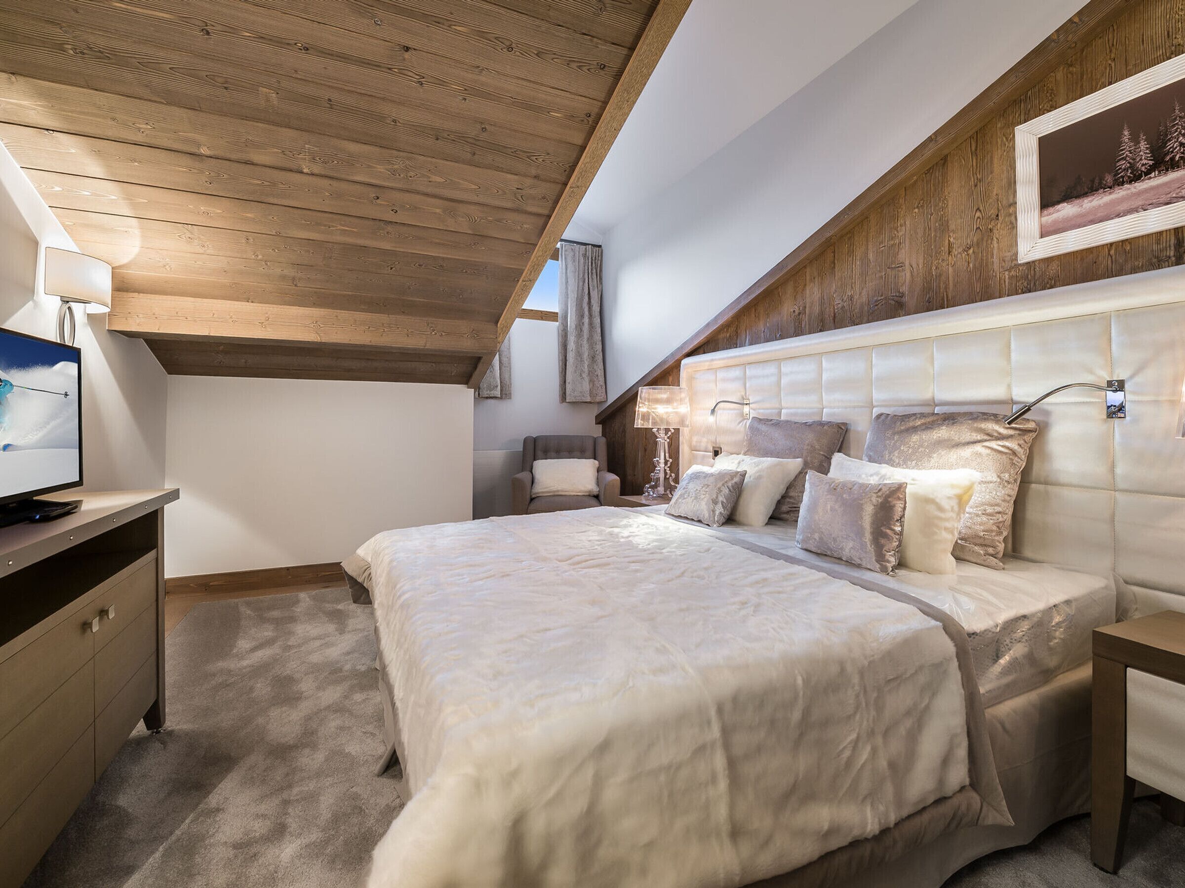 Ein Schlafzimmer mit Holzboden, einem großen Bett mit vielen Kissen, einem Nachttisch und einem Fernseher auf einem Schreibtisch.