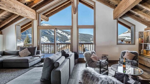 Ein Zimmer mit hohen Decken, einem großen Fenster mit Blick auf Berge, einer Couch, einem Sessel und einem kleinen Tisch mit einer Flasche und Gläsern.