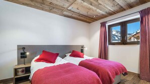 Ein Schlafzimmer mit zwei Betten, Holzboden, Holzbalkendecke, einem Fenster mit Vorhängen und einem Nachttisch mit Lampe.