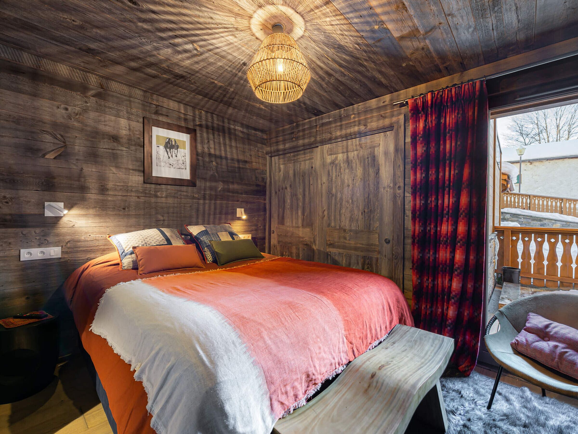 Un dormitorio acogedor con techo de madera, una cama grande con muchas almohadas, una mesita de noche, una lámpara de pared y una ventana con cortinas rojas.