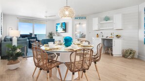 A modern living room with a large window, a dining area with wicker chairs, and a kitchen with white cabinets.
