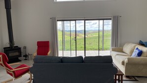 Un salon avec un canapé, un fauteuil rouge, une cheminée noire et une vue sur un paysage verdoyant, visible à travers de grandes fenêtres.