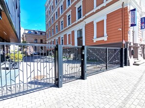 Una entrada protegida por una valla metálica, un camino pavimentado y un edificio de arquitectura clásica.