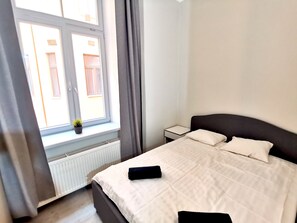 Un dormitorio con una cama, una mesita de noche, un ventanal con cortinas y un radiador.