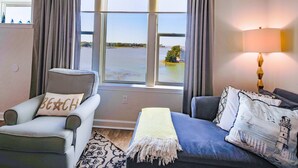 Quarto com vista para um corpo d'água, uma cadeira com travesseiro 'Beach', um sofá com almofadas estampadas e um abajur.
