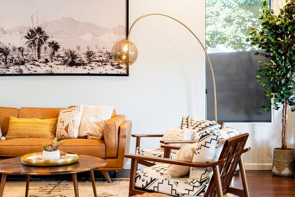 Una sala de estar con un sofá de cuero marrón, un sillón estampado, una mesa redonda de madera y una lámpara de pie adosada a la pared.
