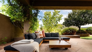 Een terras met een bank en een bijzettafeltje, omringd door groen en een heldere lucht.