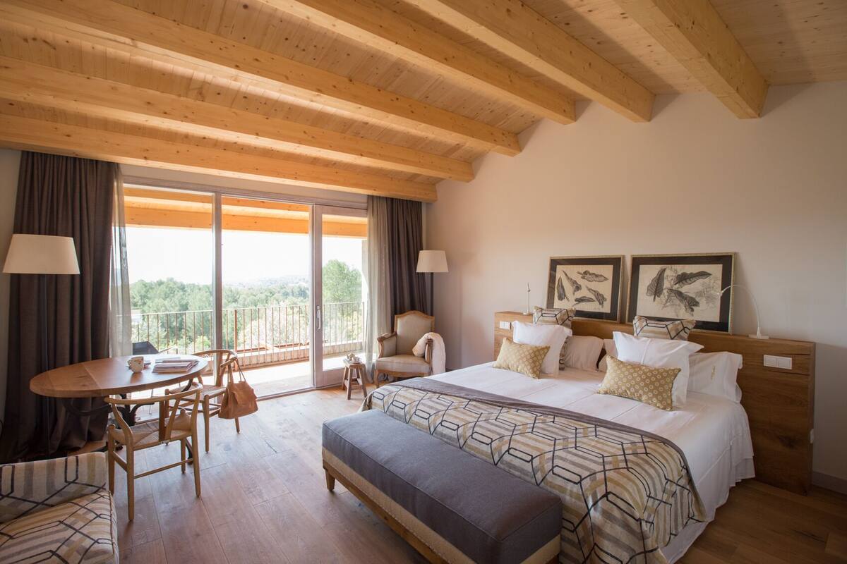 Hotelkamer met een groot bed, een klein tafeltje met stoelen, een bankje en een balkon met gordijnen.