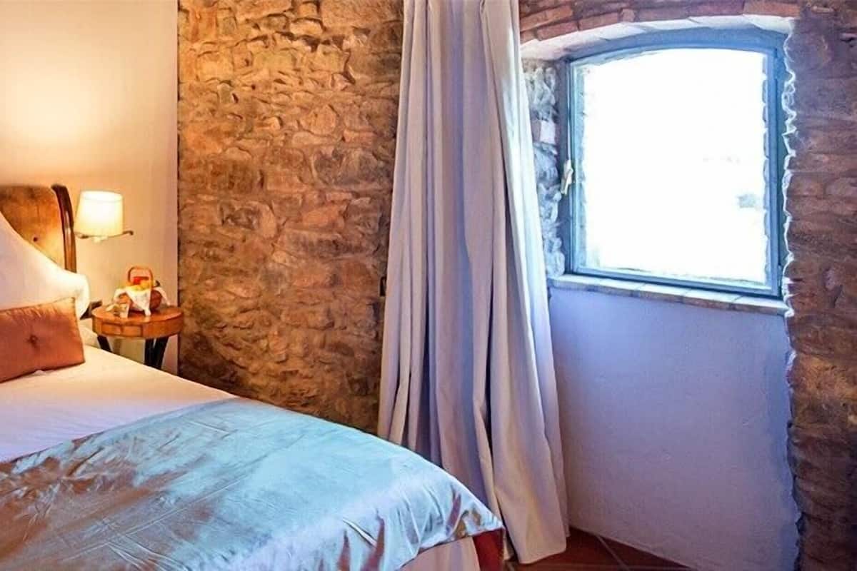 Een bed met witte lakens en twee kussens, een nachtkastje met een lamp, een stenen muur en een raam met gordijnen.