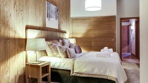 Quarto com cabeceira de madeira, mesa de cabeceira com abajur, cama com várias almofadas e uma janela grande.