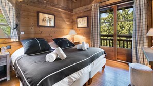 Une chambre à coucher dans une cabane en bois, avec un lit, deux tables de chevet, une petite table, une fenêtre avec des rideaux et une vue sur les arbres.