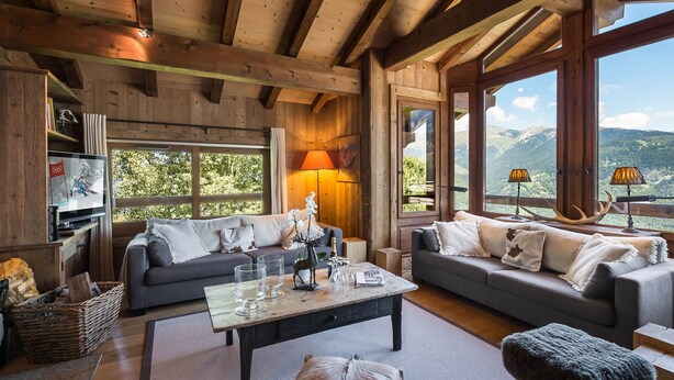 Un intérieur chaleureux de chalet en bois, avec deux canapés, une table basse et de grandes fenêtres offrant une vue sur les montagnes.