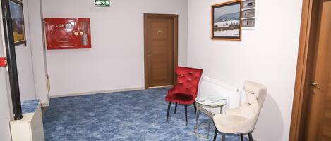 A hotel lobby with a blue carpet, a red fire extinguisher, a wooden door, and two chairs.