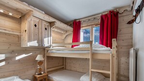 A wooden cabin room with bunk beds, a small table with a lamp, and a window with red curtains. - Bright apartment, 2 bedrooms at the foot of the slopes, pool and spa access (Méribel)