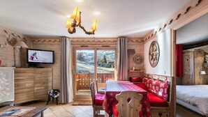 A cozy room with a wooden interior, a television, a dining table with a red tablecloth, and a view of a snowy landscape through the door. - Bright apartment, 2 bedrooms at the foot of the slopes, pool and spa access (Méribel)