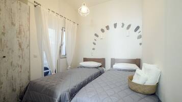 A bedroom with two beds, a window with sheer curtains, a wall-mounted light fixture, and a stone pattern on the wall.