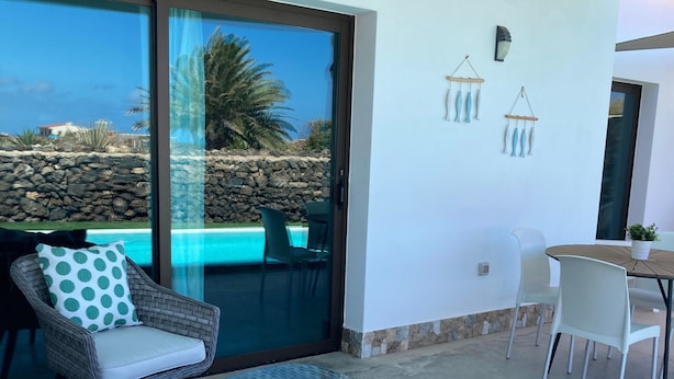 Un balcón con mobiliario de mimbre, una mesa de vidrio y vistas a una piscina y paisaje.