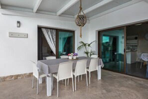 A dining area with a table set for four, white chairs, a hanging light fixture, and a view through a glass door to the kitchen.