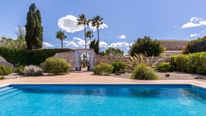 Piscina con muro y arco de piedra, rodeada de follaje y palmeras.