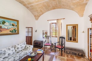 A room with a sofa, chairs, a wooden chest, a painting, a mirror, and a window with curtains.
