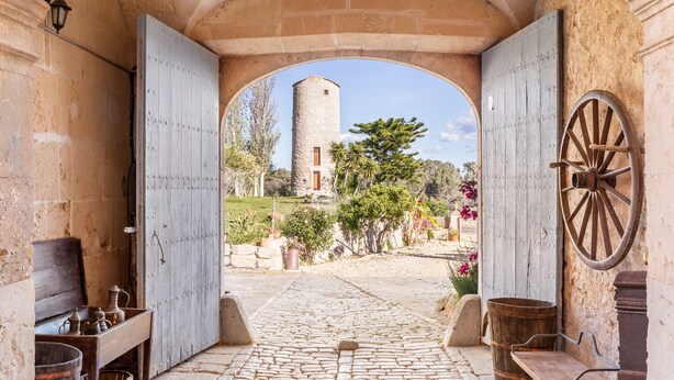 Un arco de entrada conduce a un patio con pavimento de piedra, muebles rústicos y vistas a una torre de piedra.