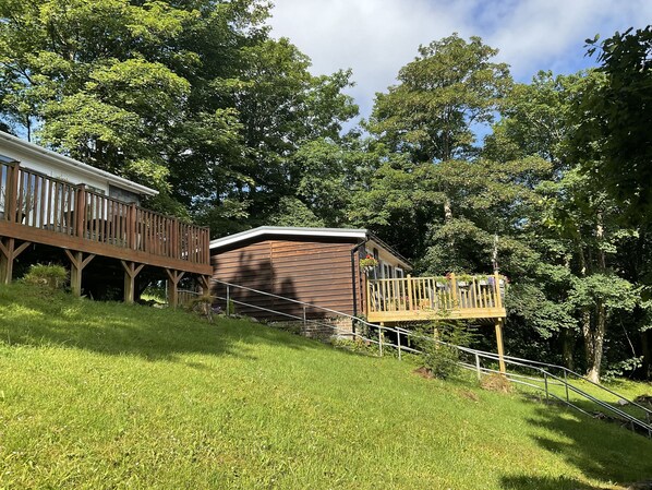 A wooden cabin with a deck and a small porch, surrounded by lush greenery and trees.