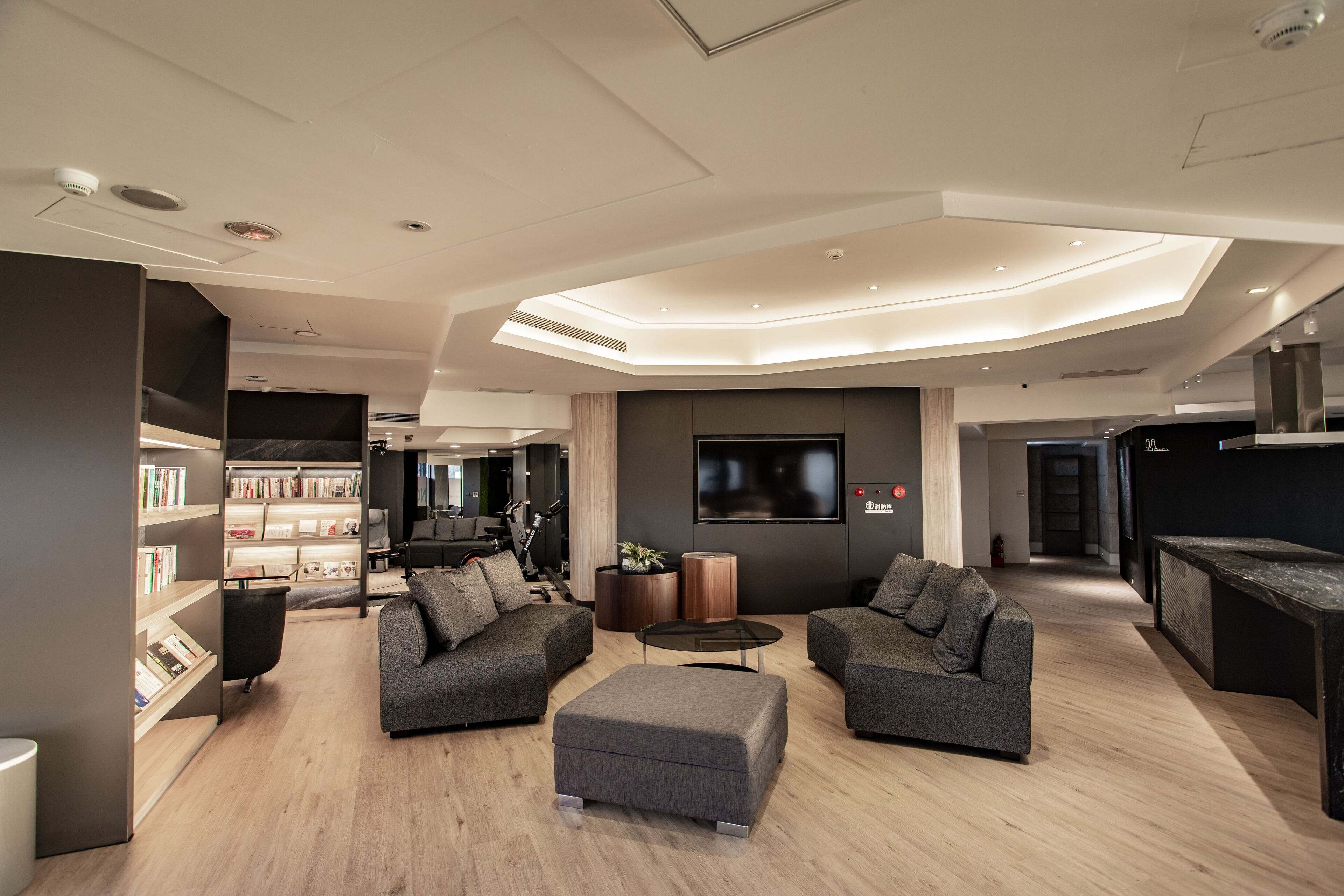 A modern living room with a central seating area, a bookshelf, and a large flat-screen TV mounted on the wall.