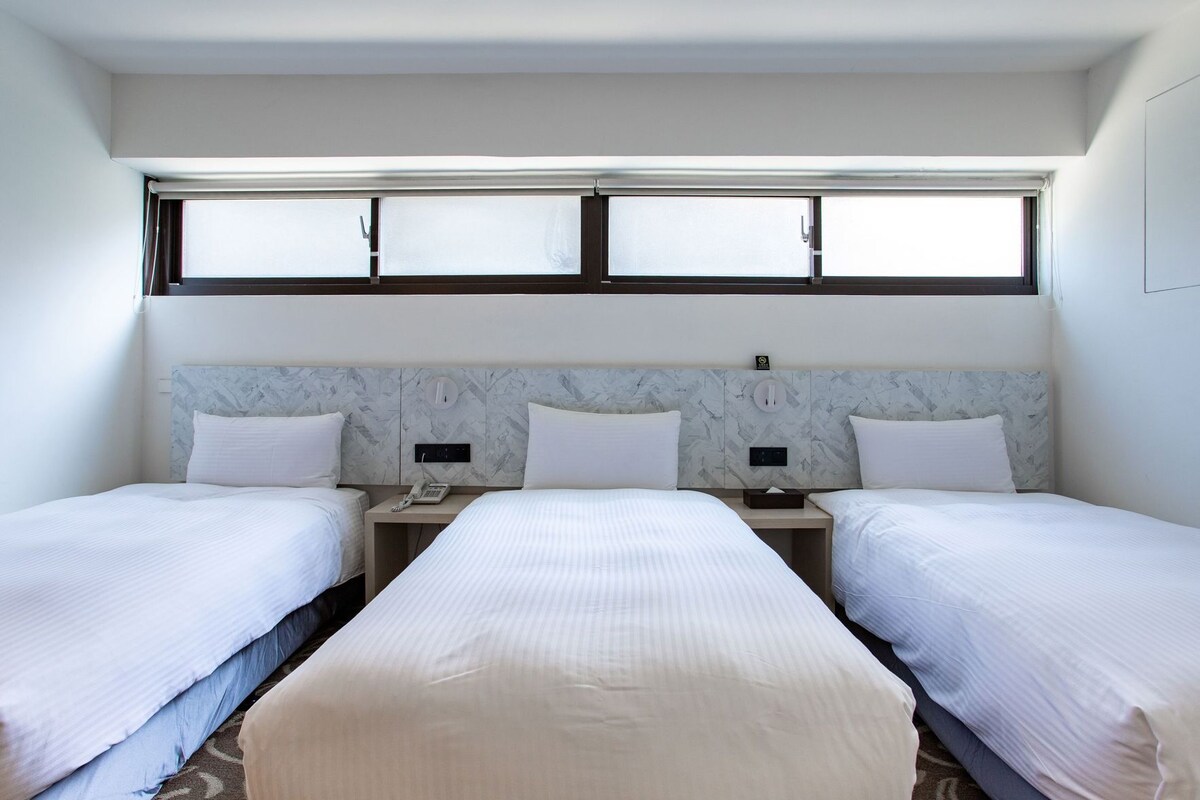 Ein Hotelzimmer mit zwei Betten, einem Marmorkopfstück, einem Fenster mit Jalousien, einem Nachttisch mit Telefon und einer Steckdose.