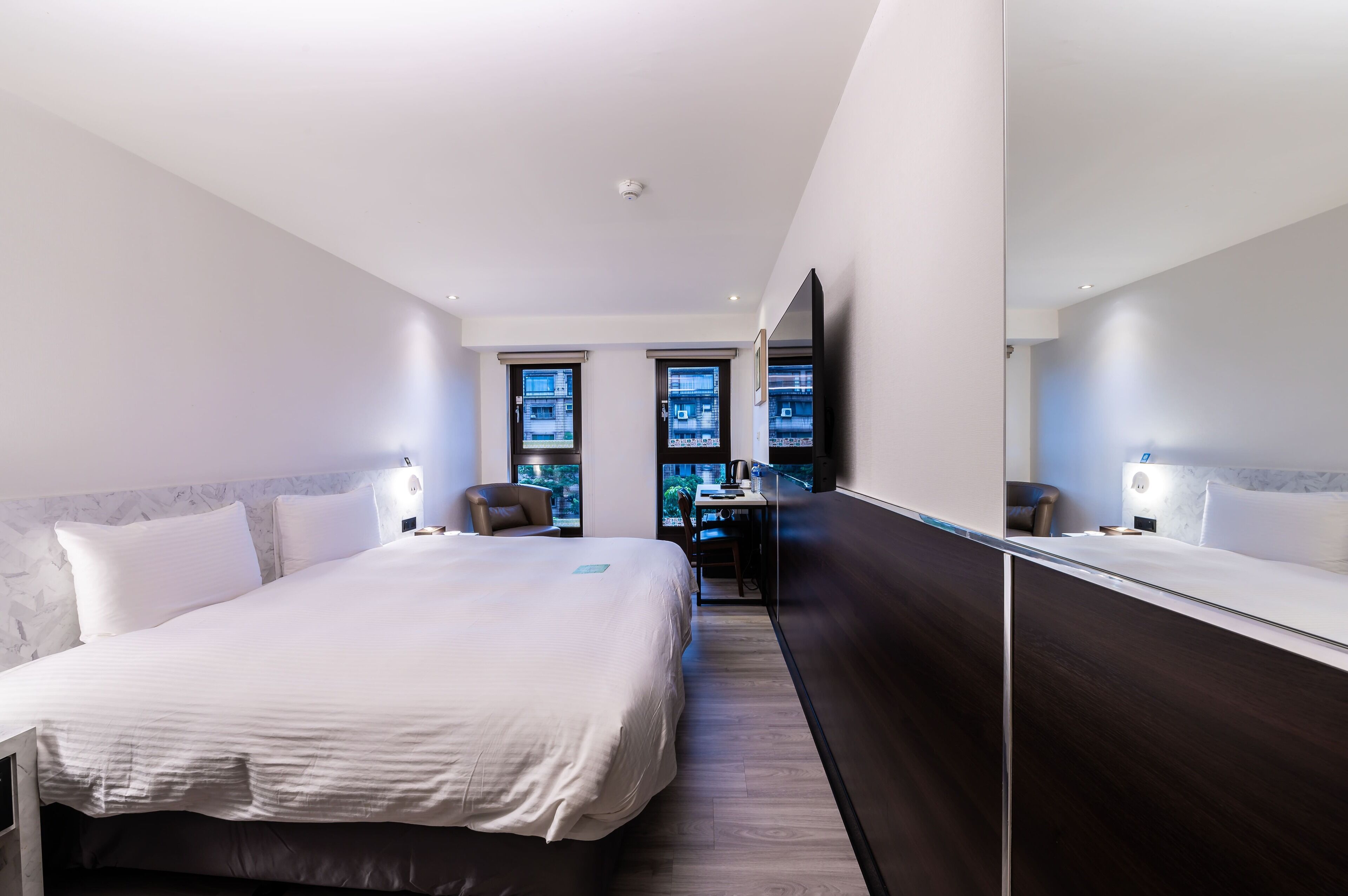 A modern hotel room with a large bed, a desk, and a view of the city through the windows.