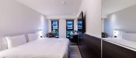 A modern hotel room with a large bed, a desk, and a view of the city through the windows.