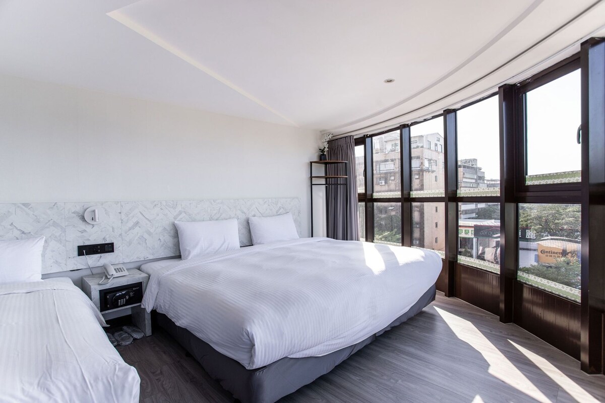 Ein modernes Hotelzimmer mit einem großen Bett, einem Nachttisch mit Telefon und Blick auf die Stadt durch große Fenster.