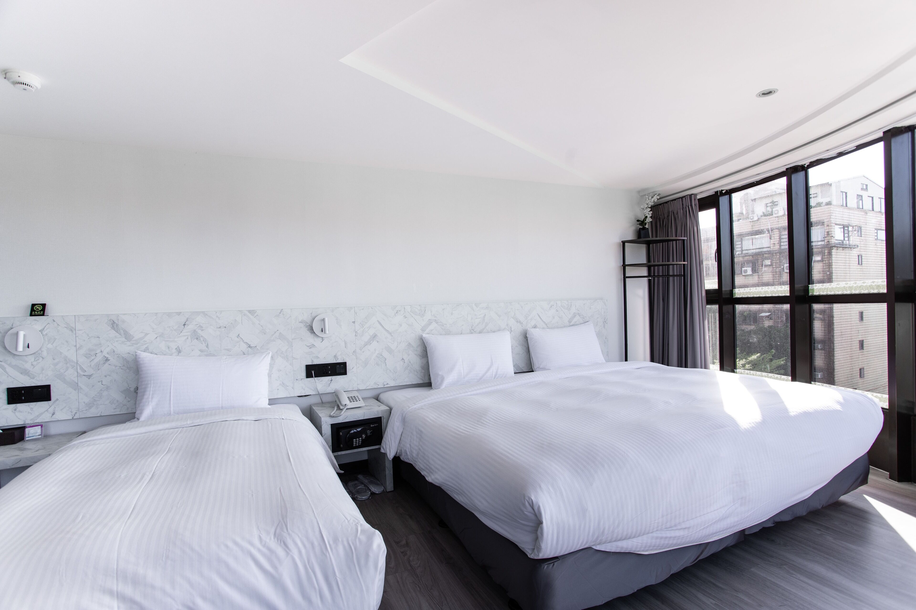 A modern hotel room with two beds, a marble headboard, a nightstand with a phone and alarm clock, and a large window with a city view.
