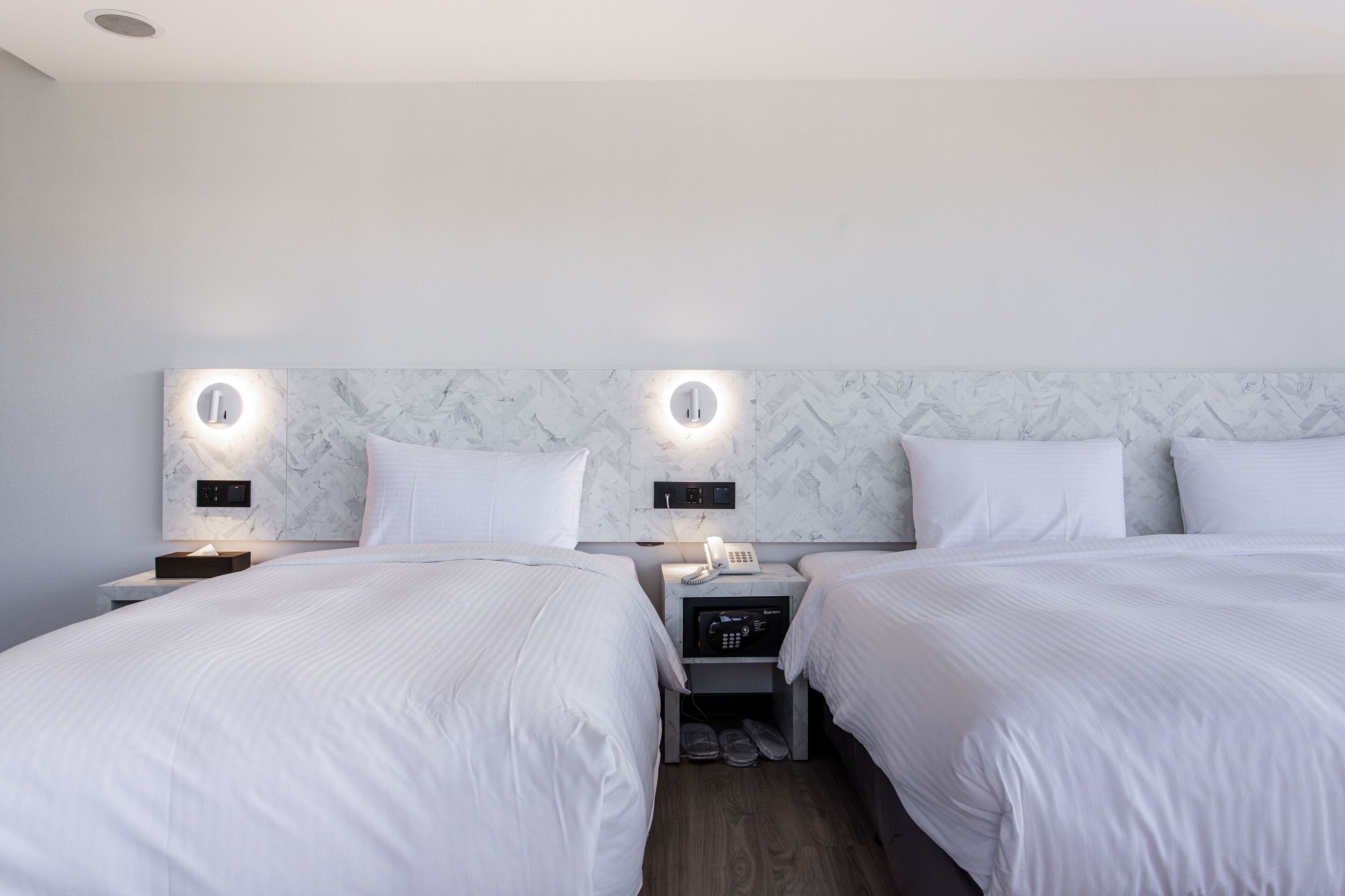 A hotel room with two beds, a marble headboard, bedside lamps, a phone, and a small storage unit.