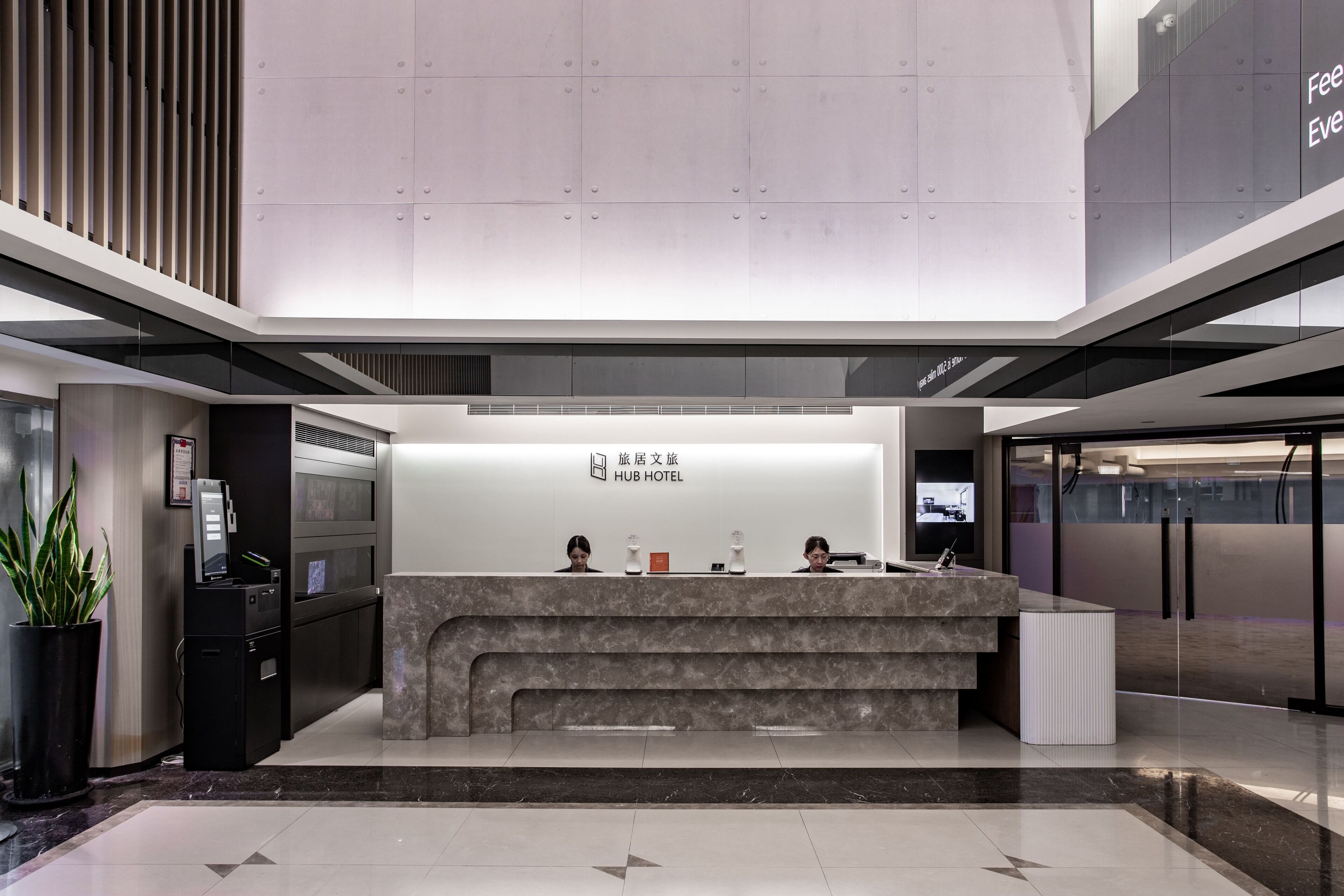 A modern hotel reception area with a marble counter, a digital kiosk, and a potted plant.
