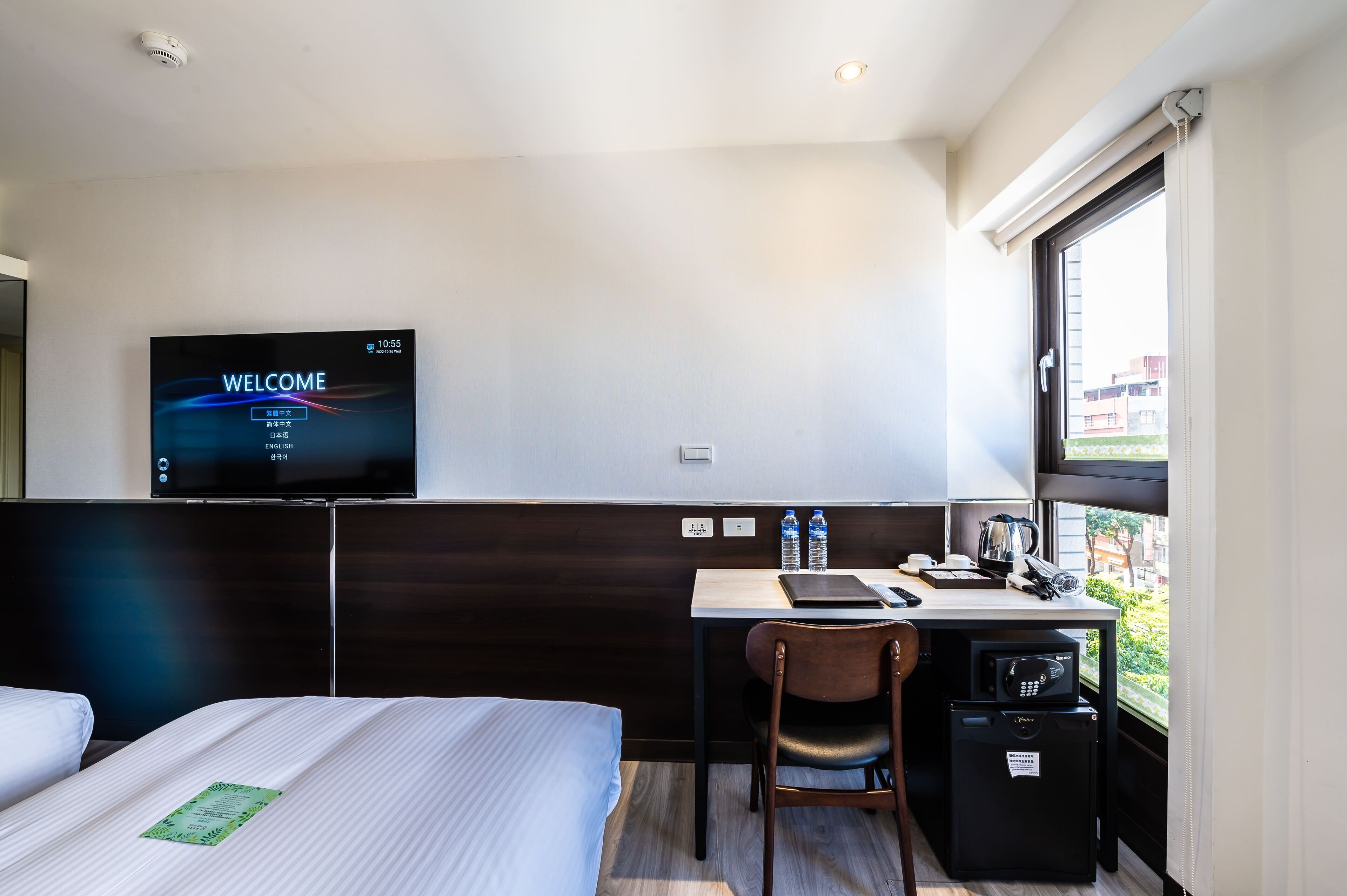A hotel room with a bed, a desk with a chair, a TV displaying "WELCOME" and language options, a window with a view, and a small refrigerator.