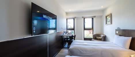 A hotel room with two beds, a desk, a chair, a TV, and a window.