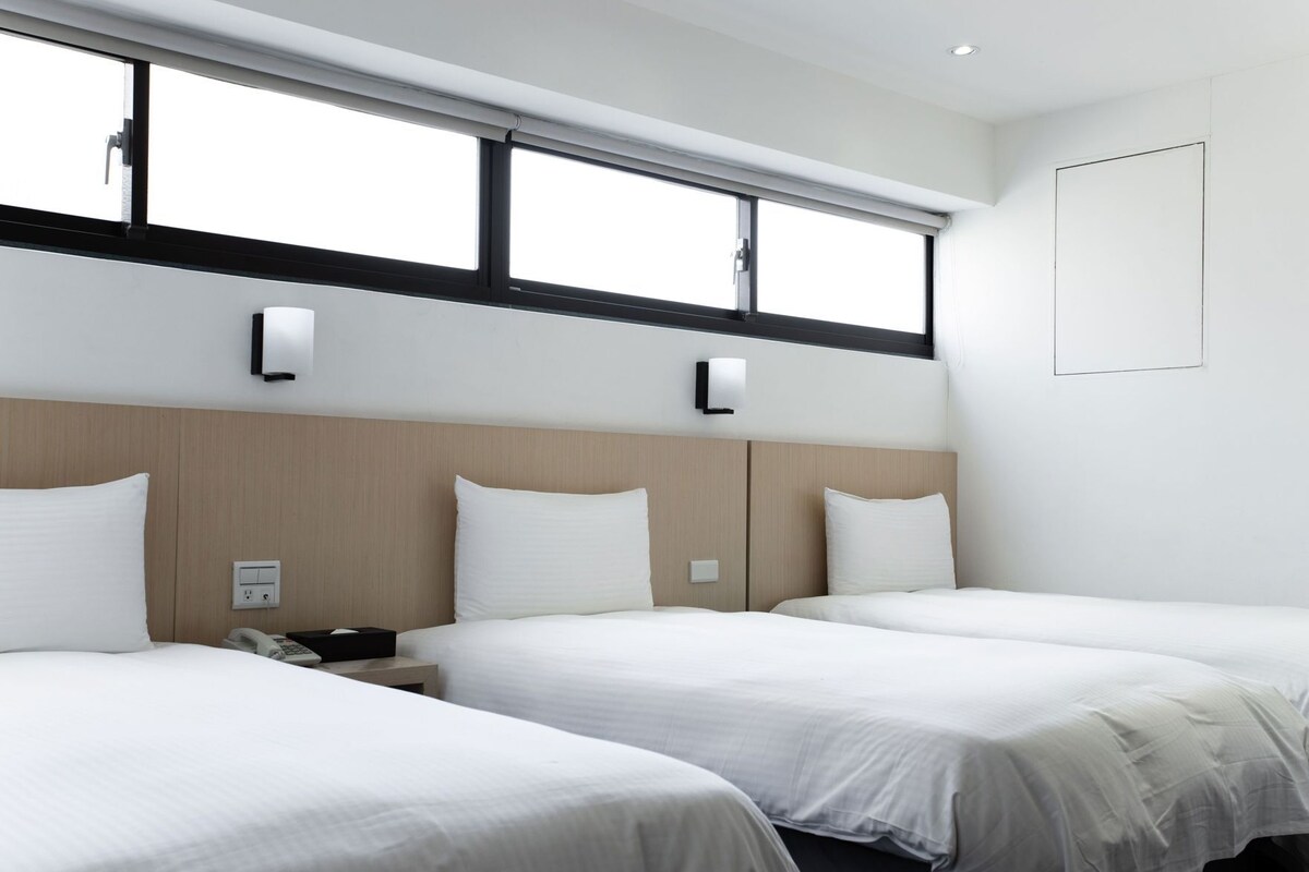 Habitación de hotel con dos camas, una ventana con persianas, una mesita de noche con teléfono y una lámpara de pared.