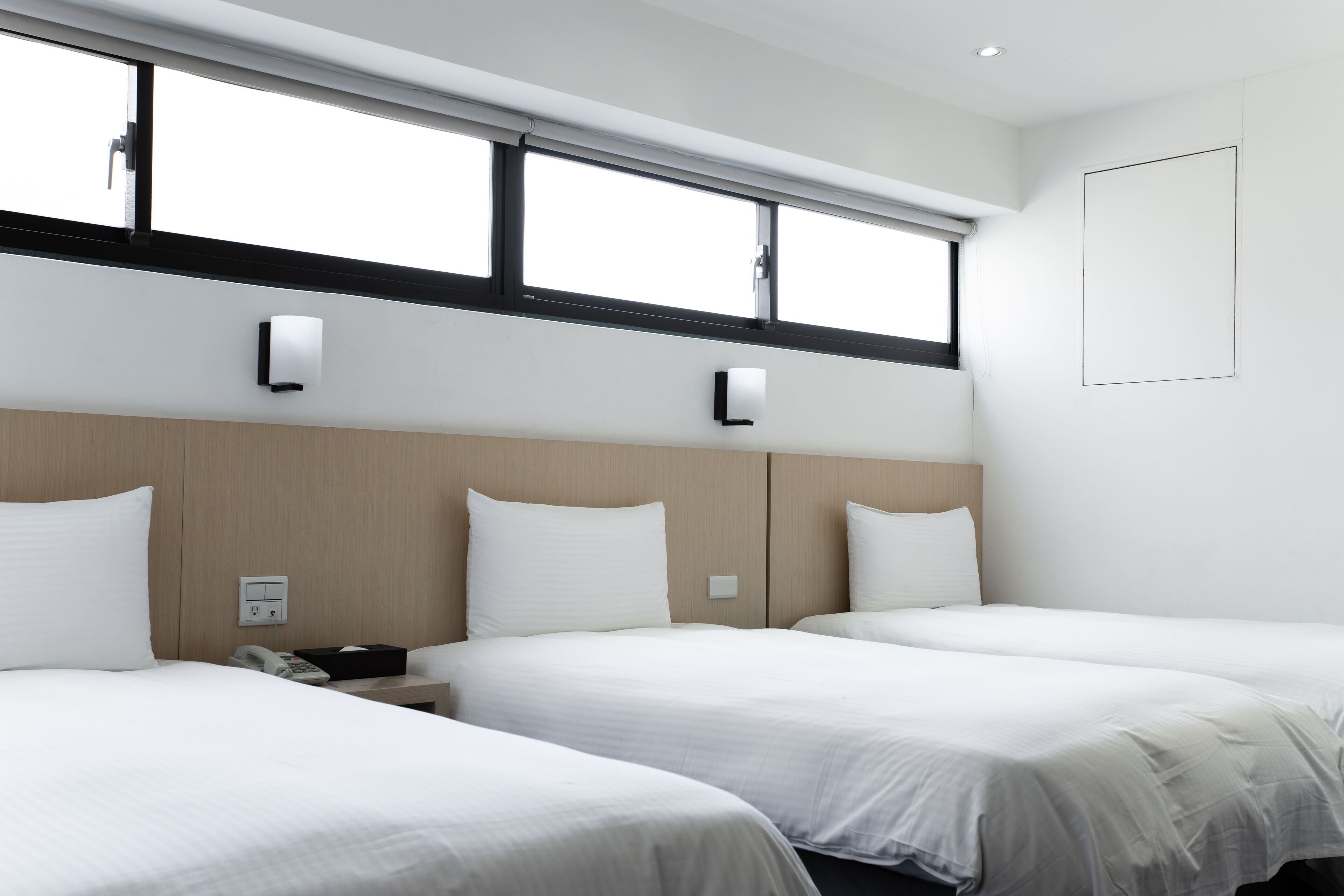 A hotel room with two beds, a wooden headboard, a window with blinds, and a wall-mounted light fixture.
