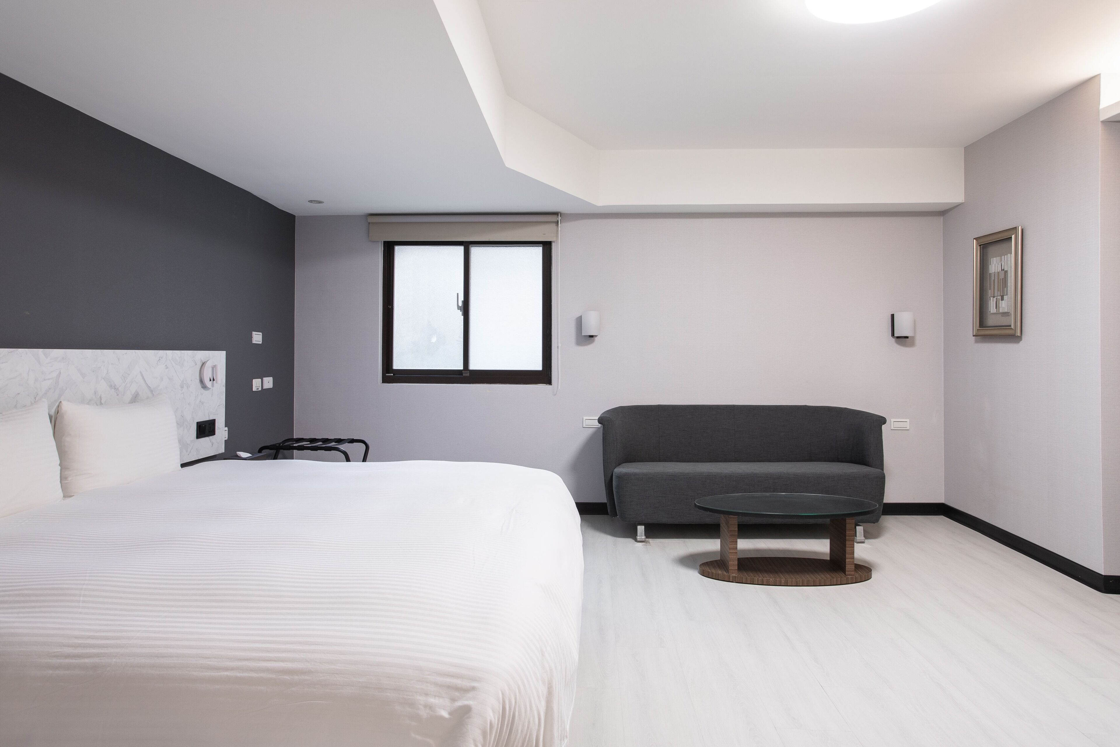 Una habitación de hotel moderna con una cama grande, un sofá gris, una mesita redonda y una ventana con persianas.