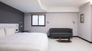 Una habitación de hotel moderna con una cama grande, un sofá gris, una mesita redonda y una ventana con persianas.