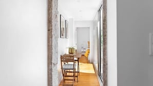 Un pasillo estrecho con una mesa de madera y sillas, una planta en maceta, y vista a una habitación con una cama y una manta amarilla.