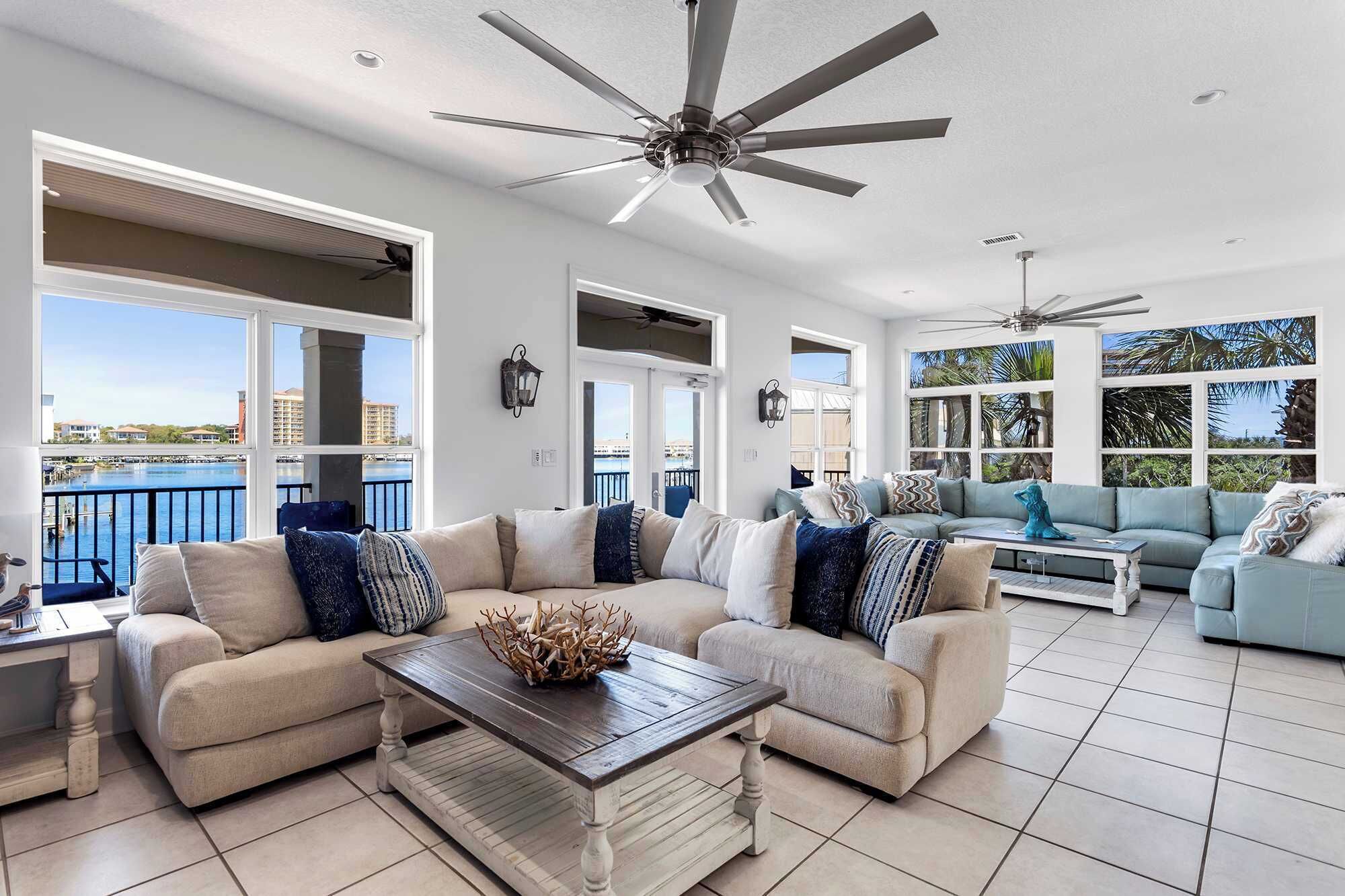 A spacious living area with a high ceiling fan, large windows offering views of a body of water, and a balcony with a railing.