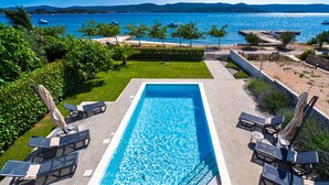 A rectangular swimming pool with a clear blue water surface, surrounded by lounge chairs and umbrellas, set against a backdrop of a beach and mountains. - Villa Roberta (Sveti Filip i Jakov)