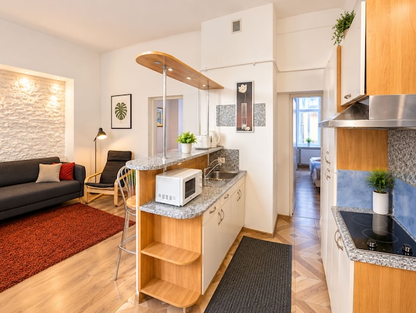 A modern kitchen with a built-in microwave, a sink, and a stove. There is a wooden chair and a small table with a plant. The kitchen is adjacent to a living area with a sofa and a lamp.